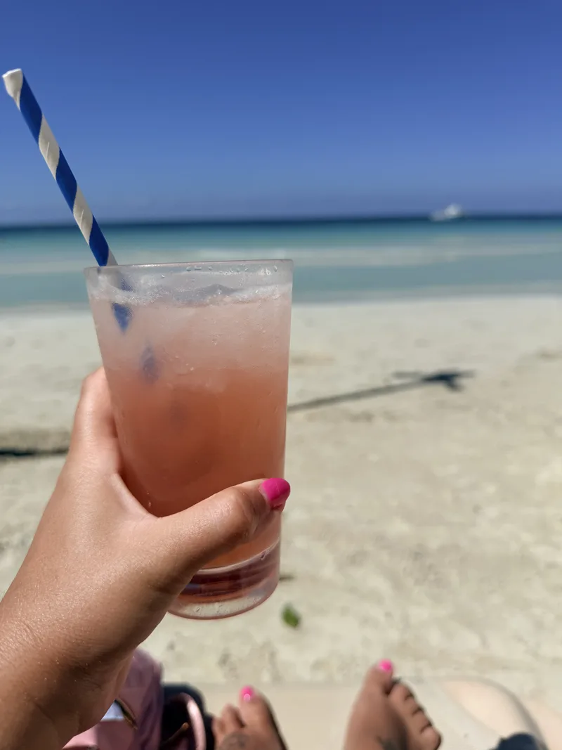 Pink drink held beside a sandy Caribbean beach with turquoise water
